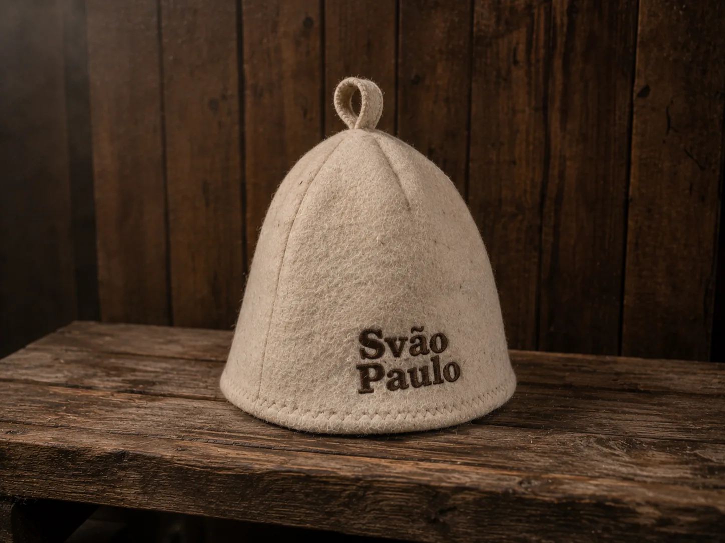 The Svão Paulo sauna cap — a cream pressed-wool felt cone with a small hanging loop on top and the wordmark 'Svão Paulo' embroidered in dark brown thread on the front. Resting on a weathered wooden bench against dark wood-paneled sauna walls.